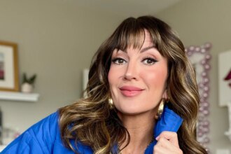 Close-up of woman with highlighted brown wavy hair and curtain bangs wearing royal blue quilted puffer jacket over blue and white striped top, holding jacket collar, layered gold charm necklace with butterfly and cross pendants, gold hoop earrings, natural glowing makeup