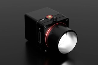 A black, box-shaped camera with a large lens and a red ring around its base sits on a reflective dark surface. The camera has a round green and red button on top and several ports on the side.