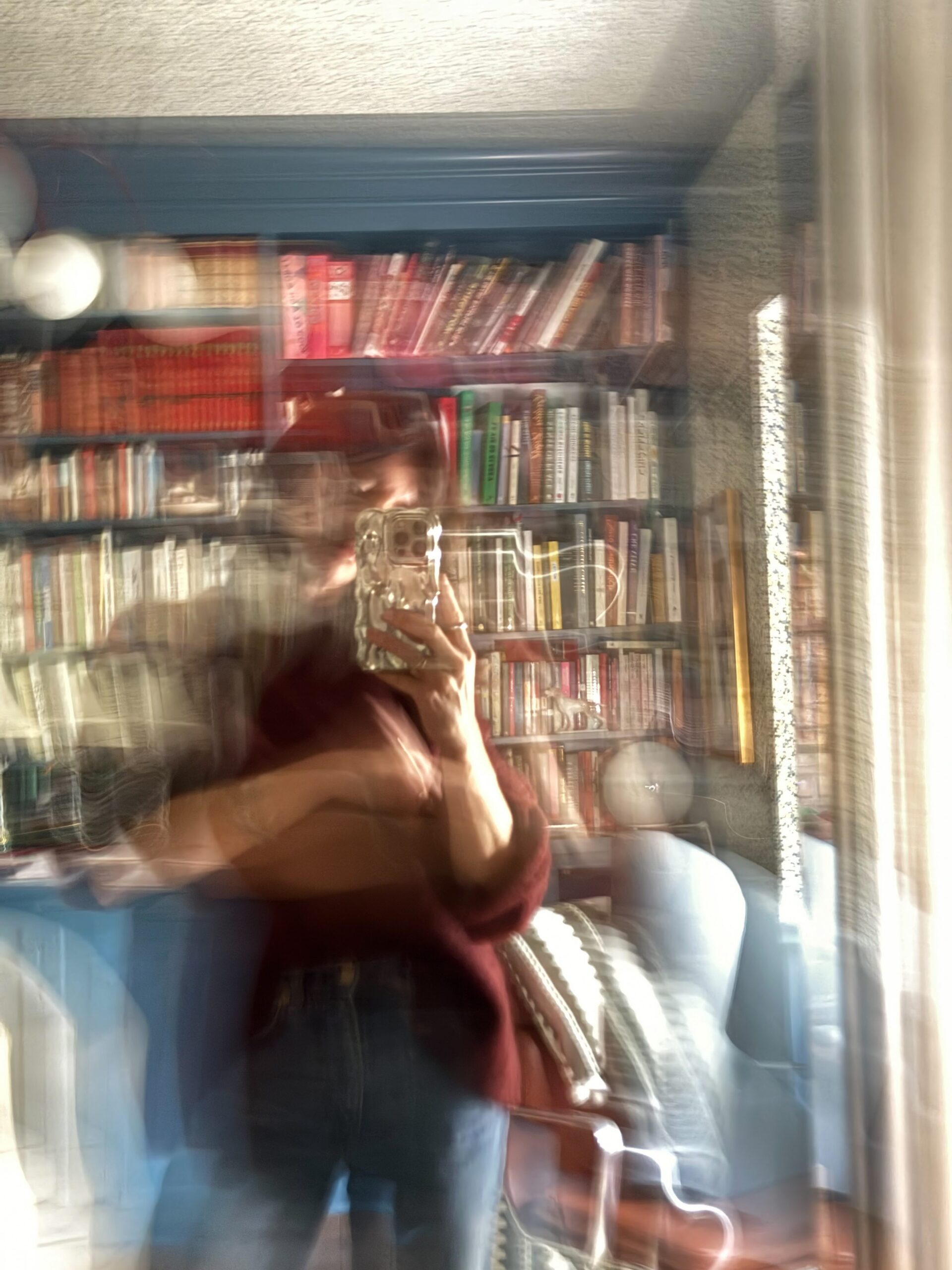 A mirror selfie of a woman in her library/home office, with a blue built-in bookshelf on one wall and blue floral wallpaper on the walls and ceiling