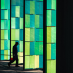 Silhouette of a man walking past vibrant green and blue glass panels, demonstrating layering and framing in photography through architectural repetition and light.