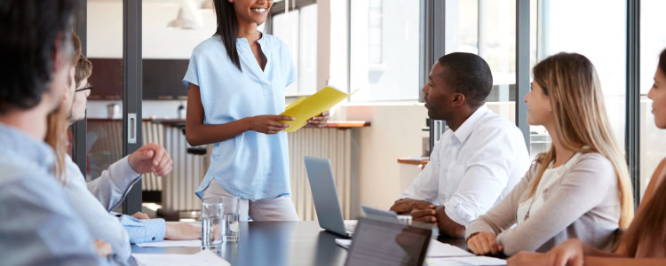 conference room with a variety of coworkers of different generations and ages and work outfits; the woman standing and addressing the group is a young Black woman wearing a blue blouse and light colored pants