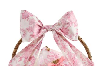 A wicker basket decorated with pink floral fabric and a large bow, filled with pastel-colored treats and Easter basket fillers, with three pastel candies placed beside it.