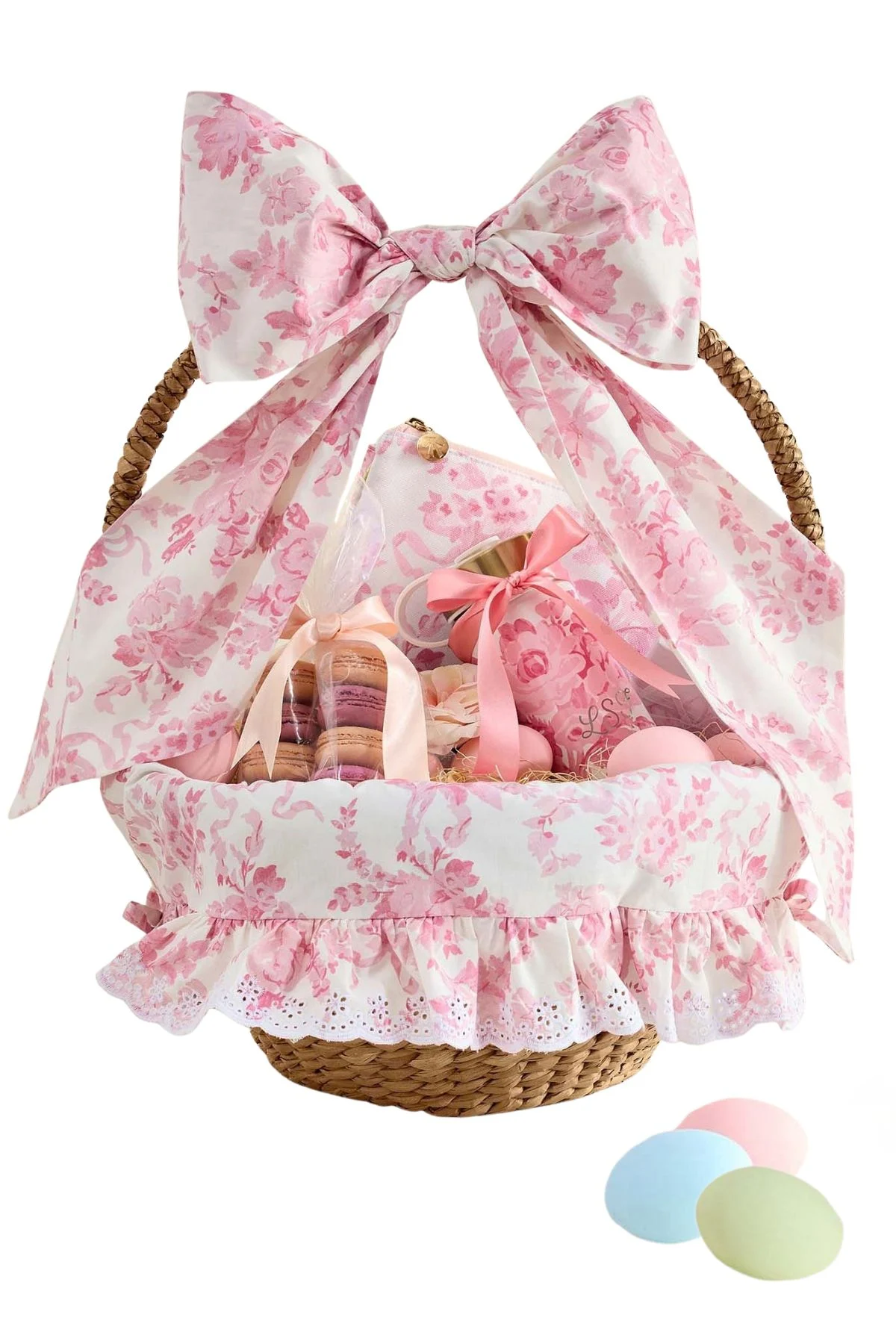 A wicker basket decorated with pink floral fabric and a large bow, filled with pastel-colored treats and Easter basket fillers, with three pastel candies placed beside it.