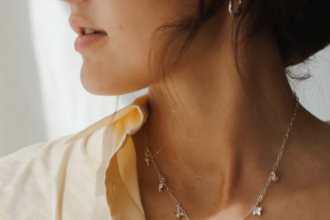 A woman wearing a cream shirt and a white ribbed top displays a silver chain necklace with small charms.