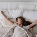 Woman wakes smiling and stretching in a bed with light-coloured bedding.