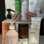 A person holds several bottles of body lotion, with additional skincare and body care products displayed on a table in front of them, all in various shapes and sizes.