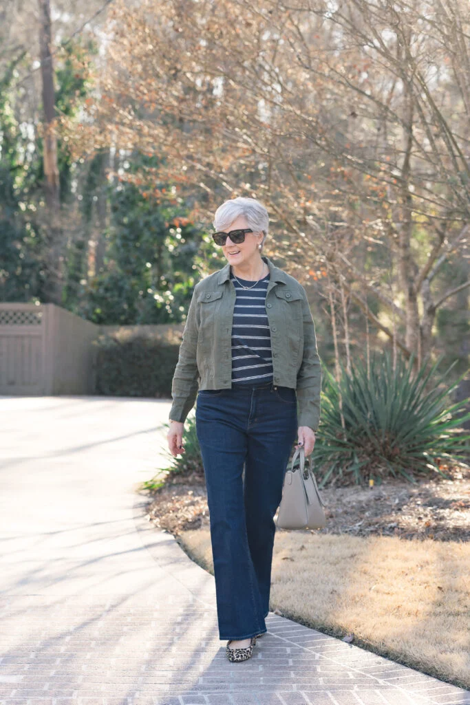 Classic style over 50 outfit with an olive utility jacket, striped top, dark jeans, and leopard flats.
