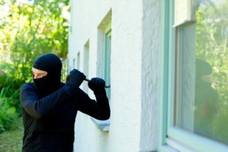 A burglar in black uses a crowbar on a white stucco window in the daylight.