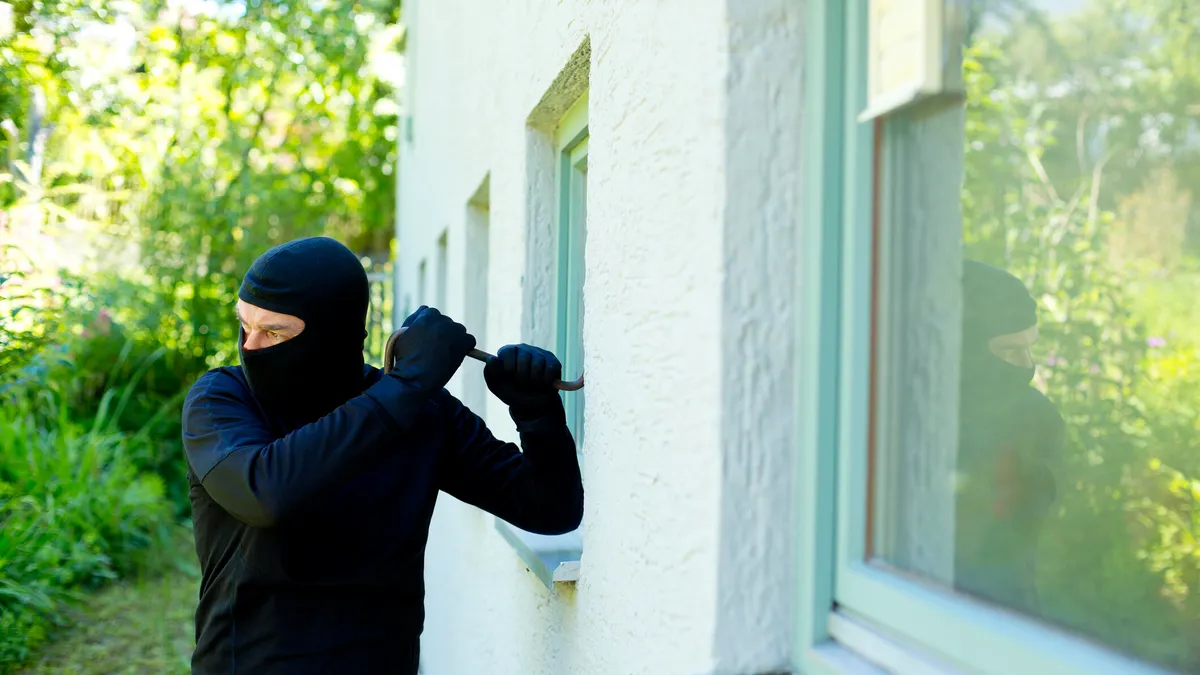 A burglar in black uses a crowbar on a white stucco window in the daylight.
