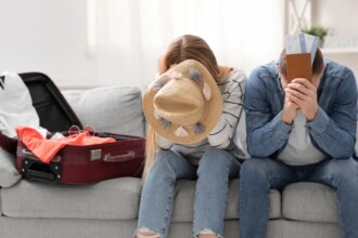 Two people sit on a sofa by a suitcase, hiding their faces with a hat and tickets.
