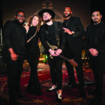 Five people dressed in black, four men and one woman, stand together on a stage in Flagler Village with musical instruments in the background. One man holds an electric guitar; all are smiling and posing to capture the rhythm of the moment. Lifestyle