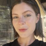 A person with short brown hair and light skin wearing a black shirt stands outside, facing the camera, with a window and yellow siding in the background.