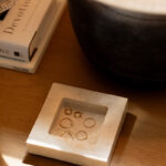 A marble tray holding several gold rings and earrings sits on a wooden surface next to a black pot and a stack of books, including one titled "Devotions.