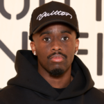 Kai Cenat wearing a black hoodie and a black cap with "Vuitton" on it stands in front of a patterned background.
