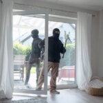 Two burglars in black enter a white room through a glass door.