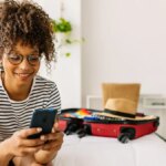 A woman sits on a white bed with a suitcase, smiling at her phone.