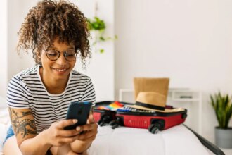 A woman sits on a white bed with a suitcase, smiling at her phone.