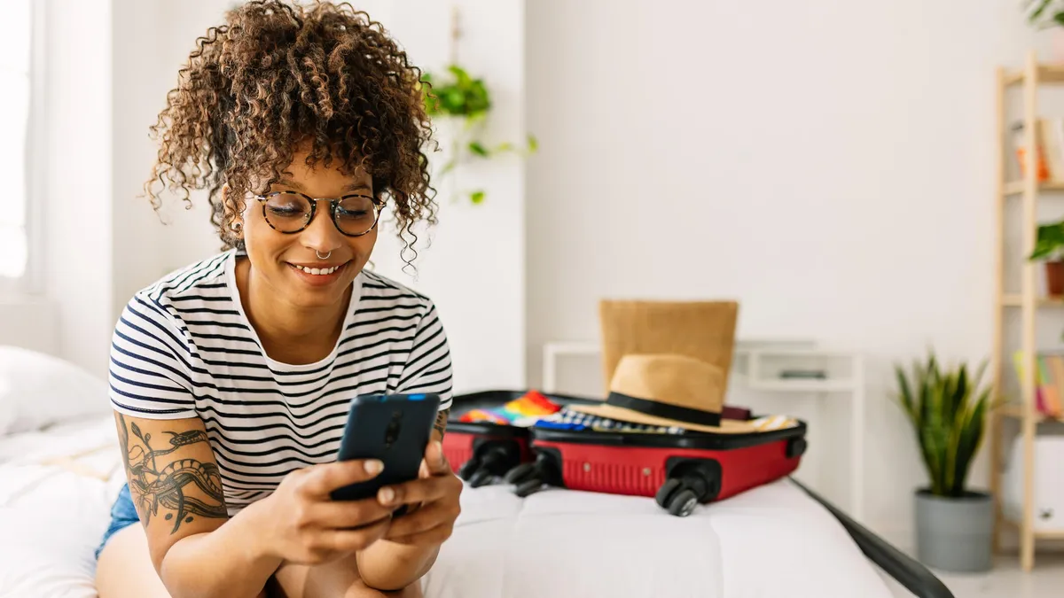 A woman sits on a white bed with a suitcase, smiling at her phone.