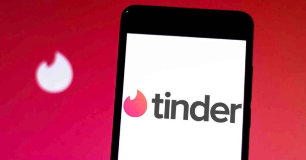 A smartphone displays the Tinder app logo on its screen, with a blurred Tinder flame icon in the background against a red gradient backdrop.