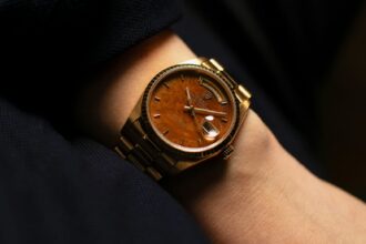 A close-up of a gold wristwatch with a brown wood-textured dial and metal bracelet, showcasing elegant wearable tech on a person's wrist against a dark fabric background. Lifestyle