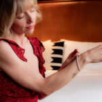 A woman in a red dress sits on a bed and fastens a bracelet on her wrist, with a pillow and framed pictures in the background.