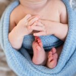 newborn swaddled in a blue blanket, holding hands together, during a posed newborn photo session