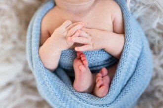 newborn swaddled in a blue blanket, holding hands together, during a posed newborn photo session