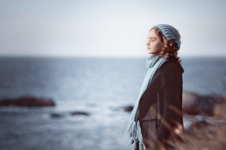 Girl in a knitted hat and scarf standing by the sea in soft light, capturing early signs of spring in nature photography with a calm coastal atmosphere.