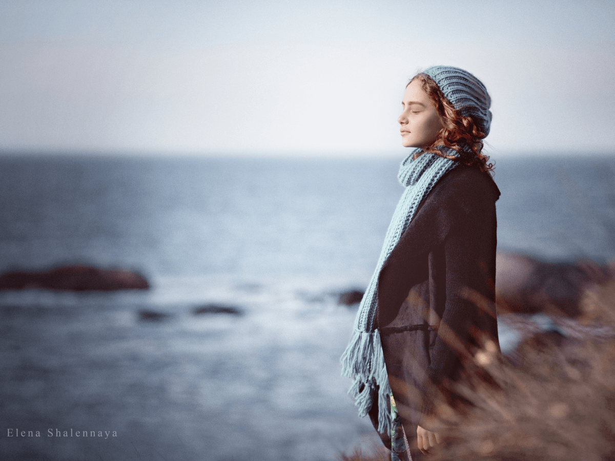 Girl in a knitted hat and scarf standing by the sea in soft light, capturing early signs of spring in nature photography with a calm coastal atmosphere.