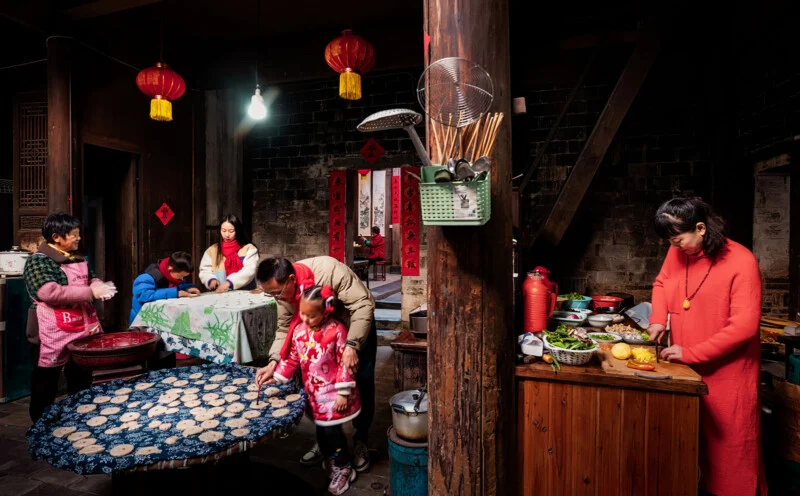 A family gathers in a warmly lit traditional kitchen, preparing food together. Children and adults work at tables with dough and ingredients, while red lanterns and festive decorations hang around the rustic room.