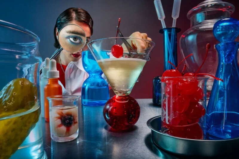 A woman wearing a white coat and holding tweezers places a cherry in a martini glass. Large glass containers and beakers with cherries, an eyeball, a pickle, and blue liquids surround her on a laboratory table.