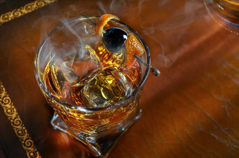 A smoked cocktail in a glass with large ice cubes, garnished with an orange twist and a dark cherry on a pick. Wisps of smoke rise from the drink, which sits on a decorative leather surface.