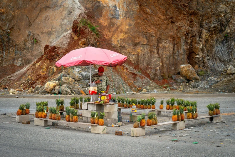 A street vendor stands under a pink umbrella selling pineapples arranged neatly on concrete blocks in a square, with a rocky hillside in the background.