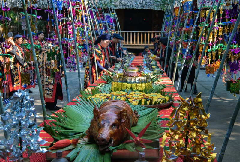 A festive, colorful table is set outdoors with a roast pig as the centerpiece, surrounded by traditional decorations, tropical leaves, and platters of food. People in vibrant cultural attire stand on either side.