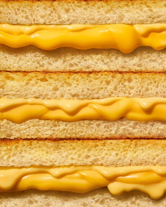 Close-up of three stacked grilled cheese sandwiches, showing golden brown toasted bread and melted yellow cheese oozing between the layers.